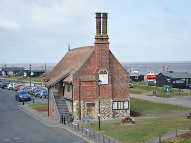 A brick building with twin chimneys near parked cars and a grassy area by the sea at Tamarisk Aldeburgh