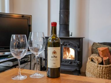 A bottle of wine and two empty wine glasses on a wooden table in front of a wood-burning stove with fire and a basket of logs in a living room at The Bothy at Snape Hall in Snape Suffolk