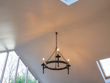 A black chandelier with four candle-like bulbs hanging from a white ceiling near large windows at The Bothy at Snape Hall in Snape Suffolk