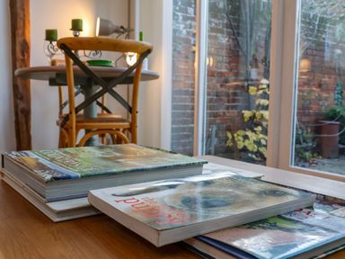 A stack of books on a wooden table with a wooden chair and a round table with green candles in the background at The Bothy at Snape Hall in Snape Suffolk