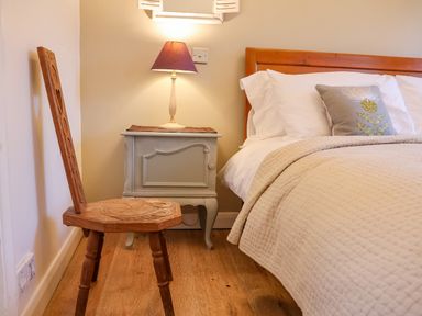 A wooden chair next to a nightstand with a lamp and a bed with white pillows and a beige quilt at The Bothy at Snape Hall in Snape Suffolk