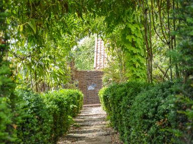 A garden pathway lined with trimmed bushes leading to a woven wooden gate with a tiled roof visible behind at The Bothy at Snape Hall in Snape Suffolk
