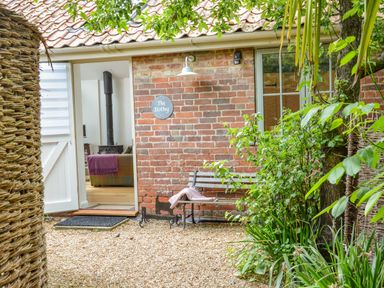 A brick building with a sign saying The Bothy an open white door revealing a room with a wood burning stove a bench with a blanket and green plants at The Bothy at Snape Hall Snape Suffolk