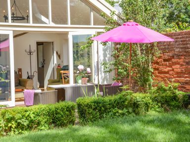 An outdoor patio with wicker furniture and a pink umbrella next to a grassy garden and a brick wall at The Bothy at Snape Hall in Snape Suffolk