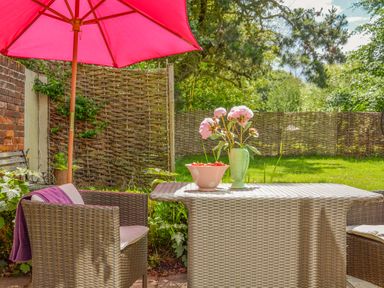 An outdoor patio with wicker chairs and table under a red umbrella with flowers in a vase and a bowl of strawberries on the table at The Bothy at Snape Hall in Snape Suffolk