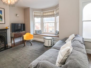 A living room with a gray sofa and white pillows a yellow chair a drum used as a table a bay window with striped blinds and patterned cushions a television on a wooden table at Osprey in Southwold