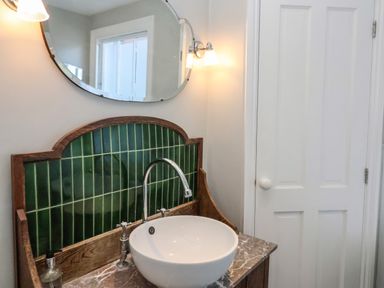 A bathroom sink with a round white basin green tiled backsplash wooden frame and a wall mirror at Osprey in Southwold