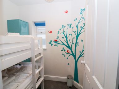 A small bedroom with white bunk beds a blue locker a window with a blue blind a wall decal of a tree with red birds and a small trash bin at Osprey in Southwold