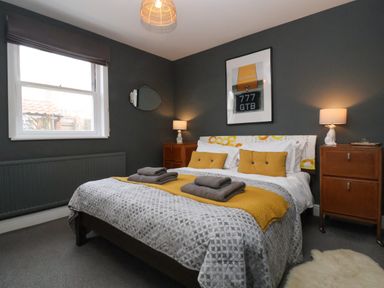 A bedroom with a double bed with gray and yellow bedding two bedside tables with lamps a window and a framed picture above the bed at Kestrel in Southwold