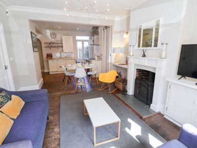 A living room with a blue sofa yellow chair white coffee table fireplace and open kitchen with dining table and chairs at Kestrel in Southwold