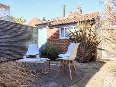 An outdoor patio area with two white chairs and a small table with cups and books surrounded by plants and a brick building at Kestrel in Southwold