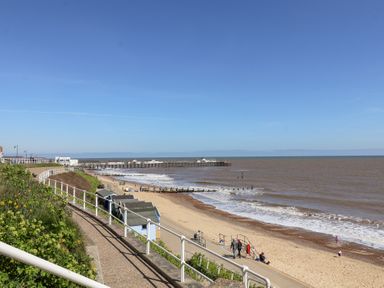 A sandy beach with a pier in the distance and people walking near small beach huts at Kestrel in Southwold