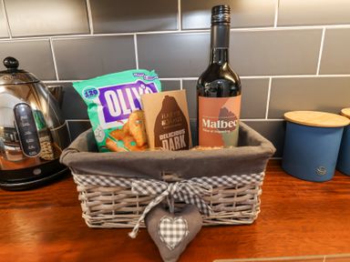 A wicker basket with snacks and a bottle of Malbec wine on a wooden kitchen counter with a kettle and blue canisters in the background at Dolphin Cottage Southwold