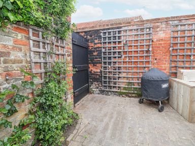 A brick patio with lattice panels on walls a covered barbecue grill and wooden seating at Dolphin Cottage Southwold
