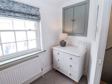A corner of a room with a white chest of drawers lamp and small cabinet under a closed wall cupboard at Dolphin Cottage Southwold