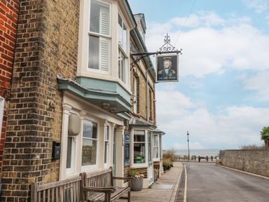 A street view of a brick building with a bench outside and a sign showing a portrait with the sea visible at the end of the road at Dolphin Cottage Southwold