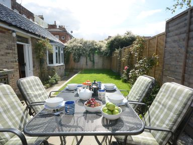 A garden patio with a table set for breakfast and cushioned chairs with a grassy yard and flowers in the background at Garden Cottage Southwold