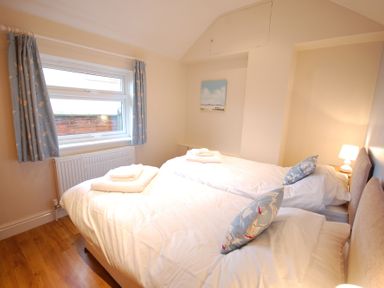 A bedroom with two single beds with white bedding and blue pillows a window with blue curtains and a bedside lamp at Garden Cottage Southwold