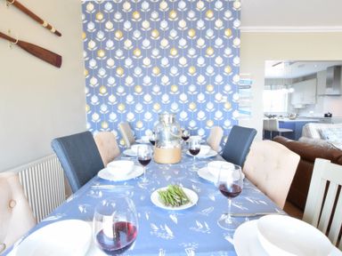 A dining table with plates wine glasses and a plate of asparagus in a room with patterned wallpaper and kitchen visible at St Marys View Southwold