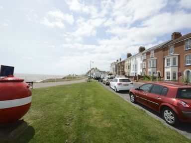 A row of houses along a street with parked cars on the right and grassy area with benches on the left near the sea at St Marys View Southwold