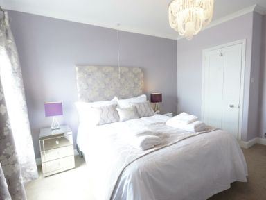A bedroom with a bed covered in white linens two bedside tables with purple lamps and a chandelier at St Marys View Southwold