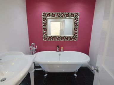 A bathroom with a white freestanding bathtub, a decorative mirror on a pink wall, and a white sink at St Marys View Southwold