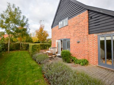 A garden with trees and bushes next to a brick house with black and gray trim at Three Chantry Barns Orford