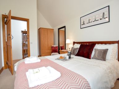 A bedroom with a bed with pillows and a tray of drinks and croissants a wardrobe a chair with red cushions a floor lamp and a large mirror at Three Chantry Barns in Orford
