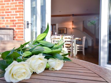 A bouquet of white roses on a wooden table with a dining area visible through glass doors at Three Chantry Barns Orford