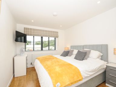 A bedroom with a large bed with gray headboard and pillows a yellow blanket bedside tables a mounted television and a window with checked blinds at Bluebell Retreat in Lostwithiel