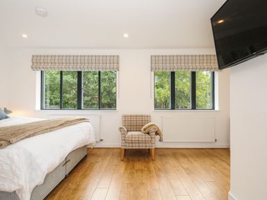 A bedroom with a bed a chair two windows and a wall mounted television at Bluebell Retreat in Lostwithiel