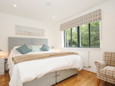A bedroom with a large bed three pillows two bedside tables with lamps a window with blinds and an armchair at Bluebell Retreat in Lostwithiel