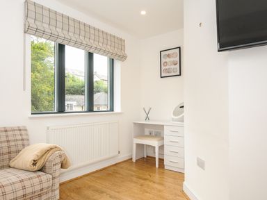 A room with a window with blinds a radiator a plaid armchair with a blanket a white desk with drawers and a stool a wall-mounted TV and framed wall art at Bluebell Retreat in Lostwithiel