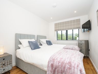 A bedroom with two single beds joined with grey headboards blue cushions white bedding pink throws a window with checked blind a wall mounted tv and two patterned bedside tables at Bluebell Retreat in Lostwithiel