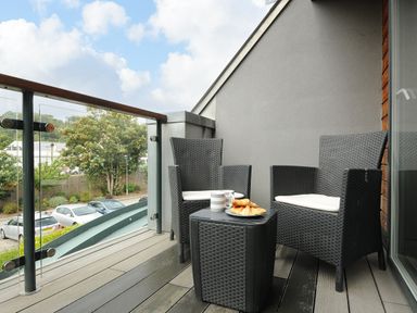 A balcony with two black woven chairs a small table with cups and croissants overlooking parked cars and trees at Bluebell Retreat in Lostwithiel