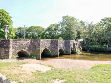 A stone arch bridge over a shallow river with trees in the background at Bluebell Retreat in Lostwithiel