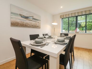 A dining room with a white table set for six near a window and a painting of a boat at Bluebell Retreat in Lostwithiel