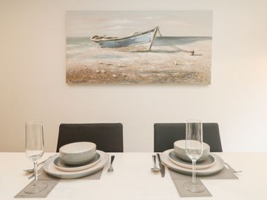 A dining table set for two with plates bowls forks knives spoons and glasses under a painting of a boat on the shore at Bluebell Retreat in Lostwithiel