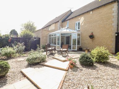 A garden patio with wooden chairs and a glass conservatory attached to a stone house at Oakmead in Moreton-In-Marsh