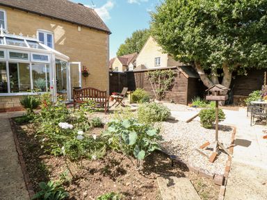 A garden with a wooden bench a chair a birdhouse plants and a wooden shed at Oakmead in Moreton-In-Marsh