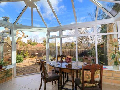 A dining area with a wooden table and chairs inside a glass conservatory overlooking a garden at Oakmead in Moreton-In-Marsh