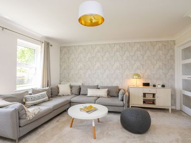 A living room with a gray sectional sofa a white round coffee table a gray pouf a sideboard with a lamp and small plants at Bryntirion in Moelfre