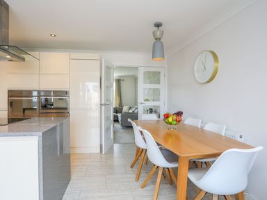 A kitchen with wooden dining table white chairs fruit bowl wall clock and open door leading to living room at Bryntirion in Moelfre