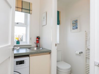 A small utility room with a sink washing machine and window next to a bathroom with a toilet towel rail and framed picture at Bryntirion in Moelfre