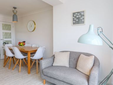 A dining area with a wooden table and white chairs and a small grey sofa with cushions and a floor lamp at Bryntirion in Moelfre
