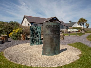 An outdoor memorial with bronze plaques and a building with lifeboats signs in the background at Bryntirion in Moelfre
