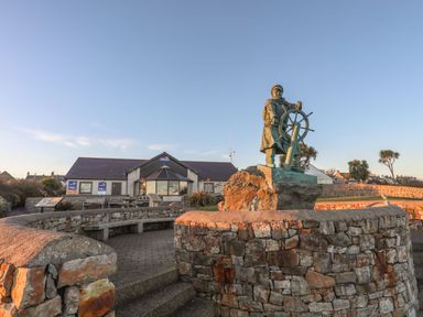 A statue of a person steering a ship's wheel in front of a building with rnlI lifeboats signs at Bryntirion in Moelfre