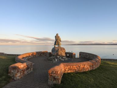 A stone statue on a pedestal surrounded by a curved stone wall near a body of water at Bryntirion in Moelfre