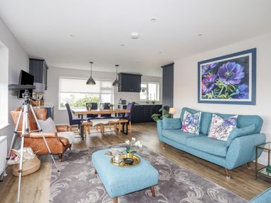 A living room and kitchen area with a blue sofa and ottoman a brown leather armchair a dining table with chairs and bench a telescope and floral painting at Watlands Mynytho near Abersoch