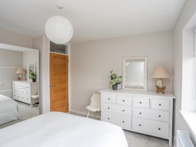 A bedroom with a white dresser a mirror a lamp a plant a wooden door a white chair and a bed at Watlands in Mynytho near Abersoch